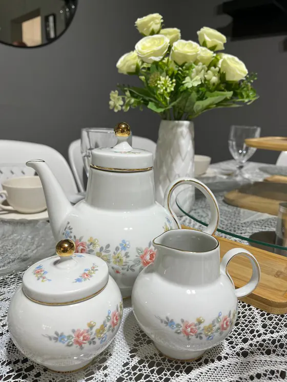 Juego de té de porcelana blanca con flores y detalles dorados, junto a un florero con rosas blancas