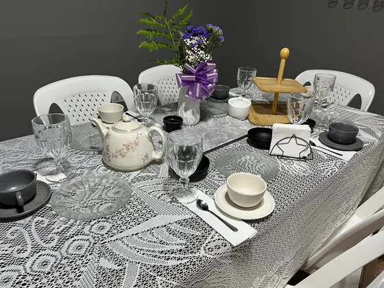 Mesa con mantel de encaje, tetera con flores rosadas, florero con moño lila y tazas grises y crema